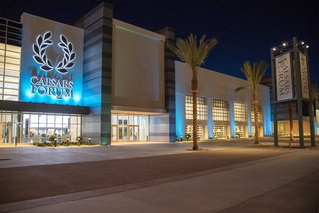 Exterior view of Caesars Forum convention center at night, with illuminated signage and palm trees along the entrance walkway.