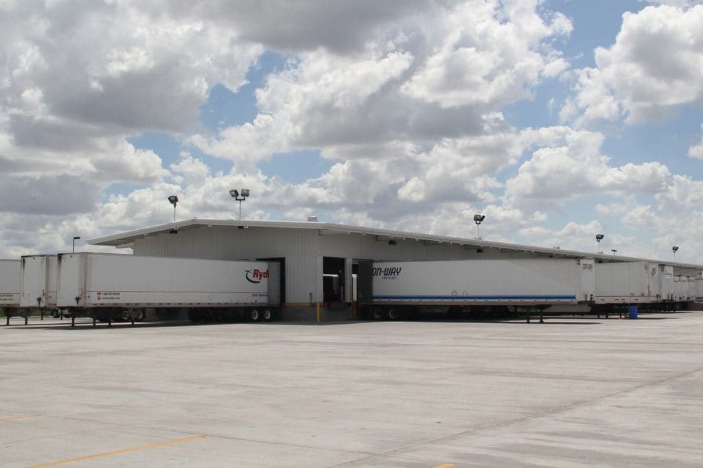 A large warehouse with multiple semi-trailers parked at loading docks under a partly cloudy sky.