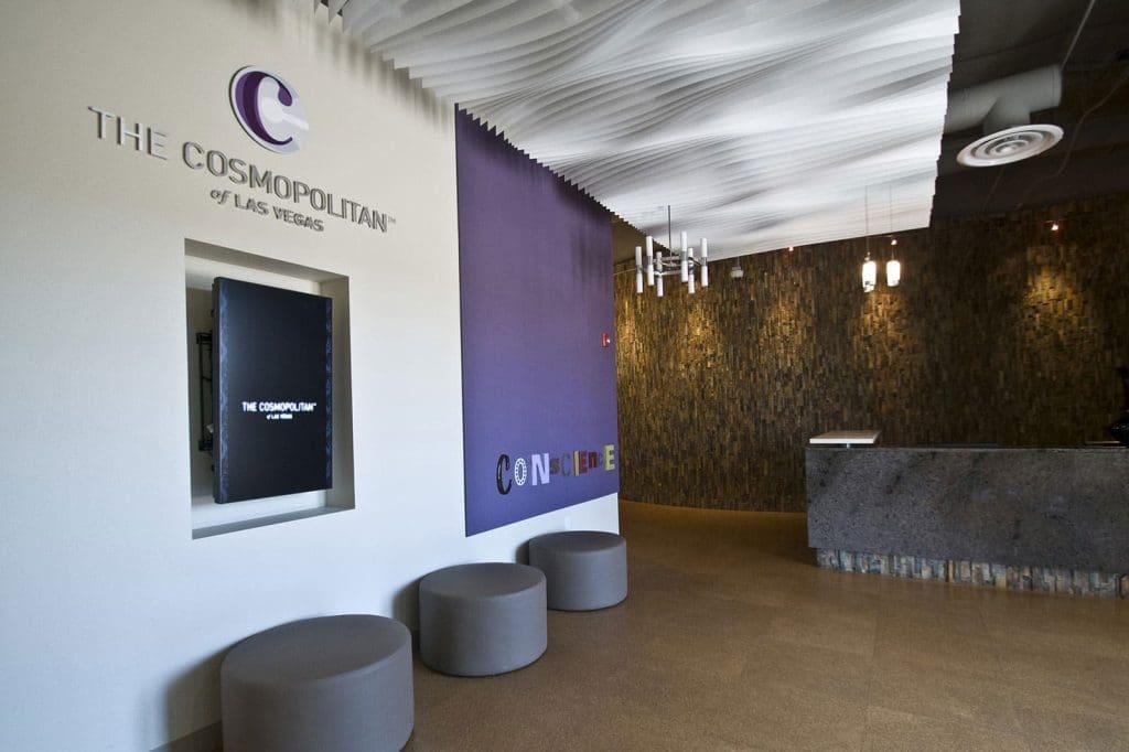 Lobby area of The Cosmopolitan of Las Vegas with a purple accent wall, round gray seats, a digital display, and a modern reception desk.