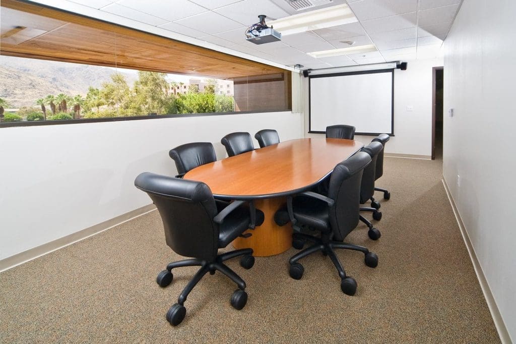 A conference room with a long wooden table, eight black office chairs, a projector, a projection screen, and a large window with a view of trees and mountains.