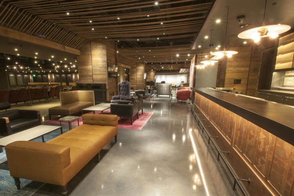 Spacious modern lounge with wood accents, upholstered seating, a long bar, polished concrete floor, and warm overhead lighting.