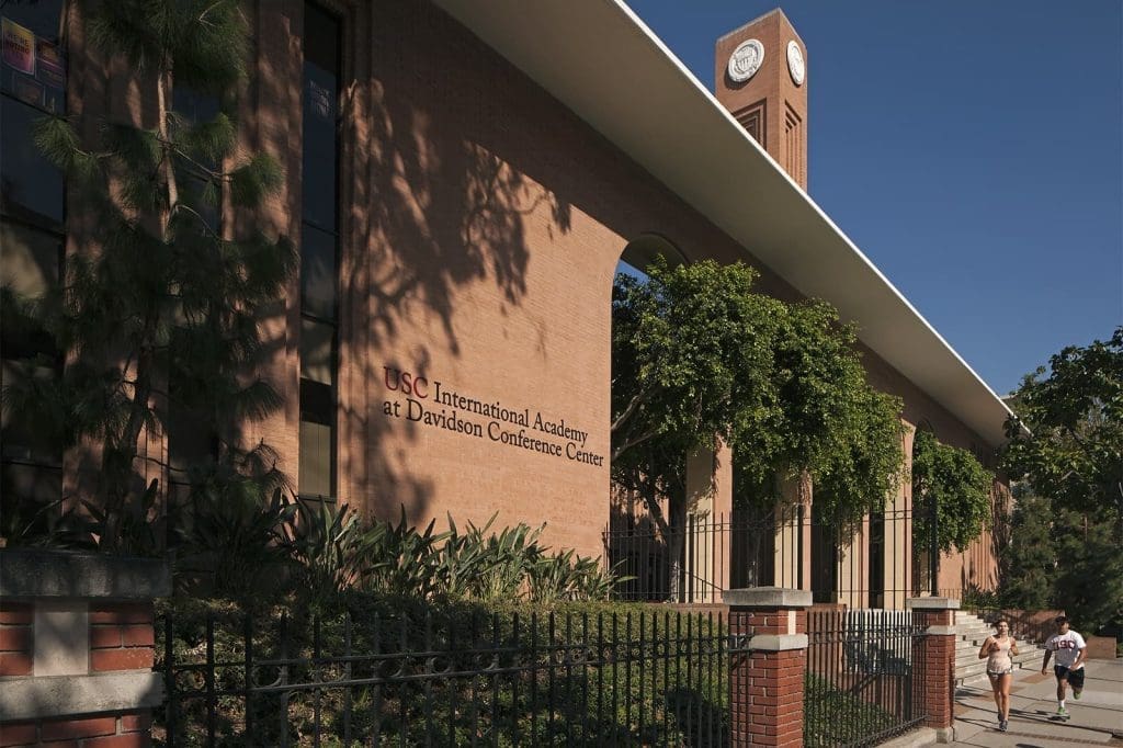 Red brick building with "USC International Academy at Davidson Conference Center" on the facade, clock tower in the background, trees and two people walking on the sidewalk.