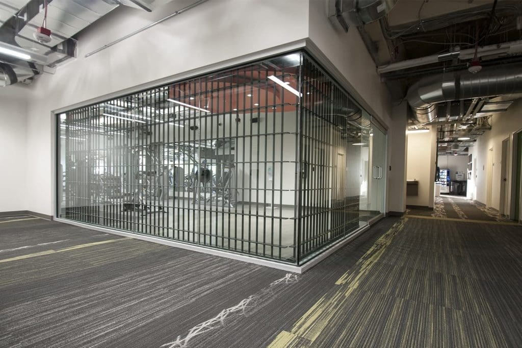 A modern glass-walled gym room with metal bars viewed from a hallway with exposed ceilings and patterned carpet.